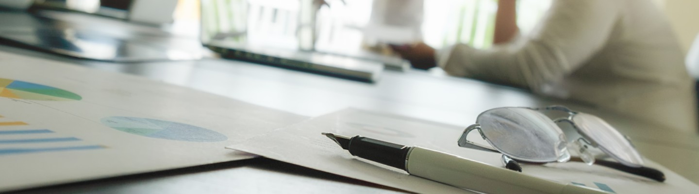 office desk with closeup of a pen and pair of glasses on top of papers