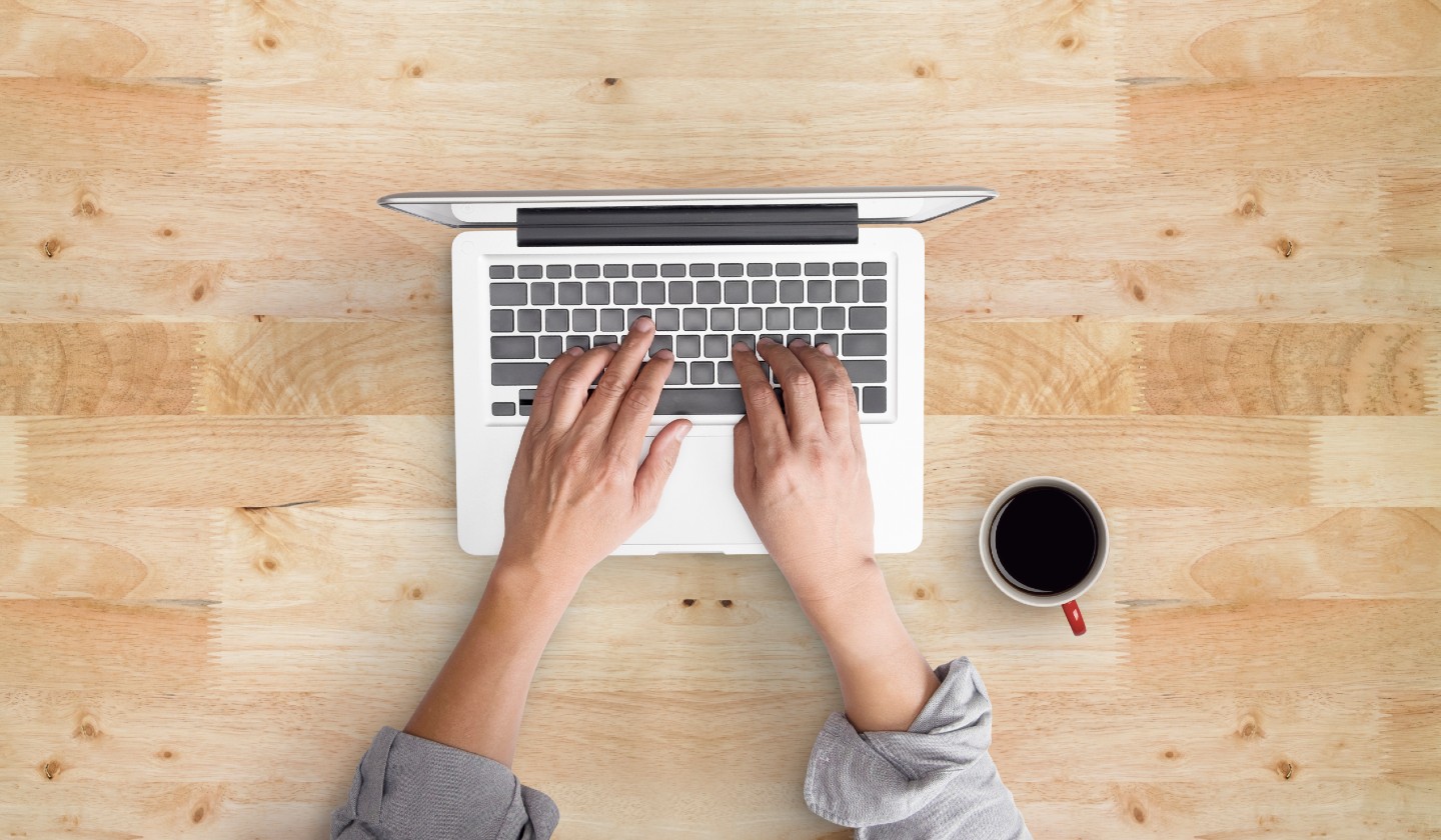 looking down on two hands typing laptop on a wooden desk with a mug of black coffee on the right side of the laptop