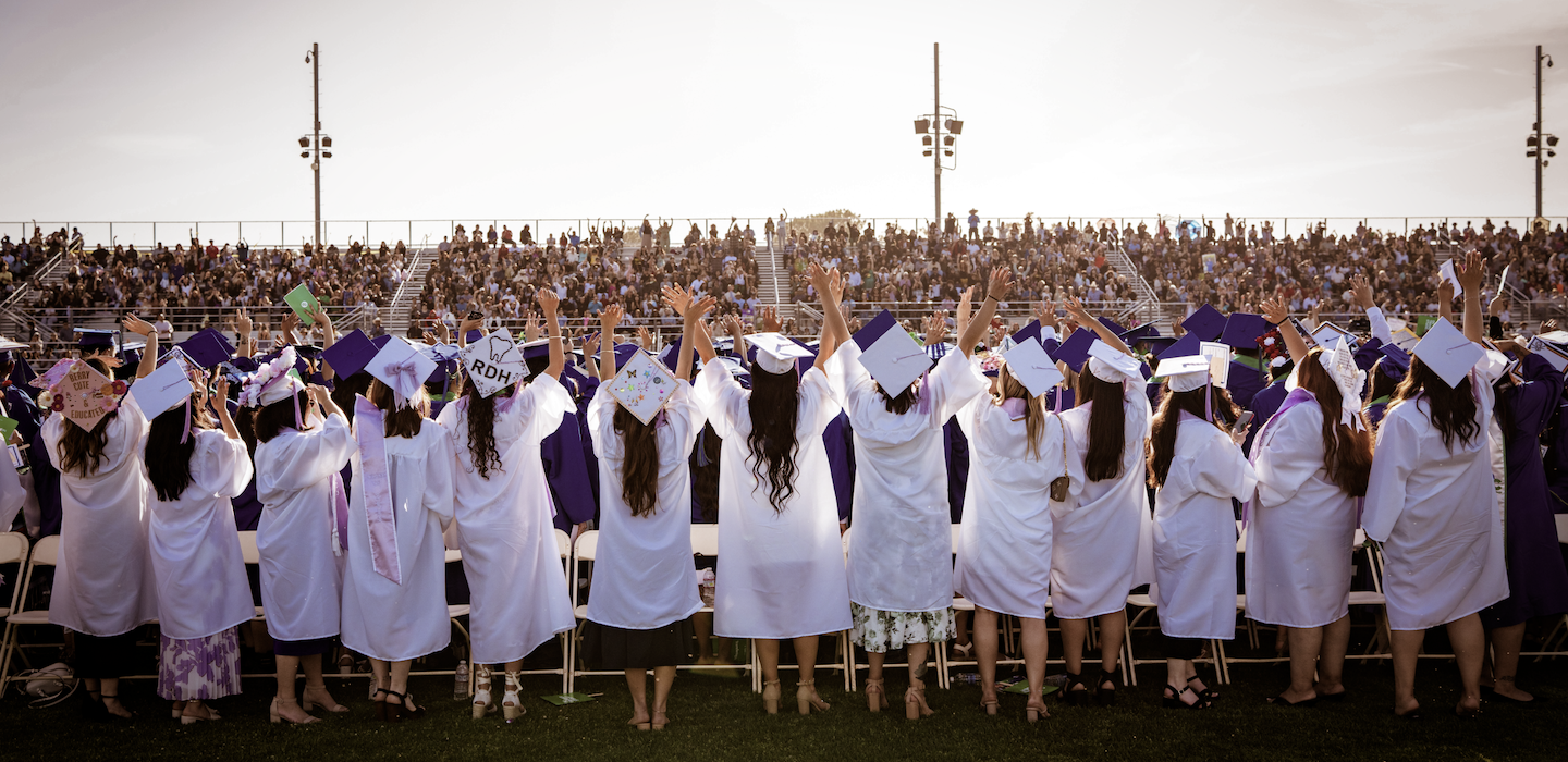 Graduates in caps and gowns raise their hands in celebration at a packed outdoor commencement ceremony.