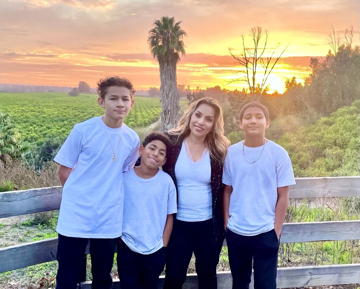 Michelle Castelo standing with three grandsons wearing white shirts and black pants in front of a nature scene with a sunset