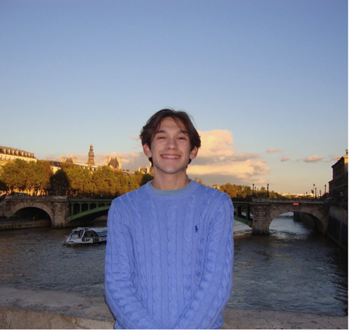 "A young man in a light blue sweater smiles while standing by a river with bridges in the background."