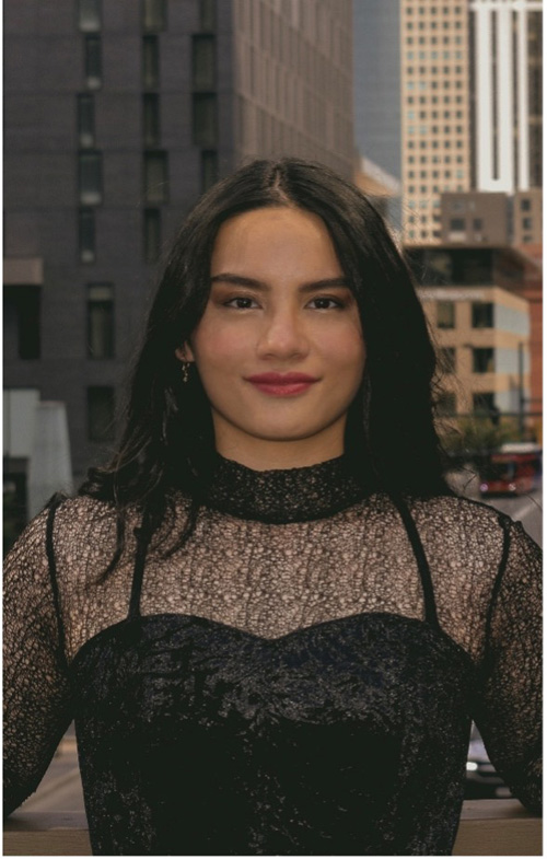 **"A woman with long dark hair wears a black lace top and poses in front of a cityscape."**