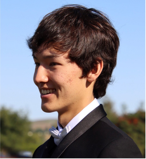 "A young man in a black tuxedo and bow tie smiles while looking to the side."