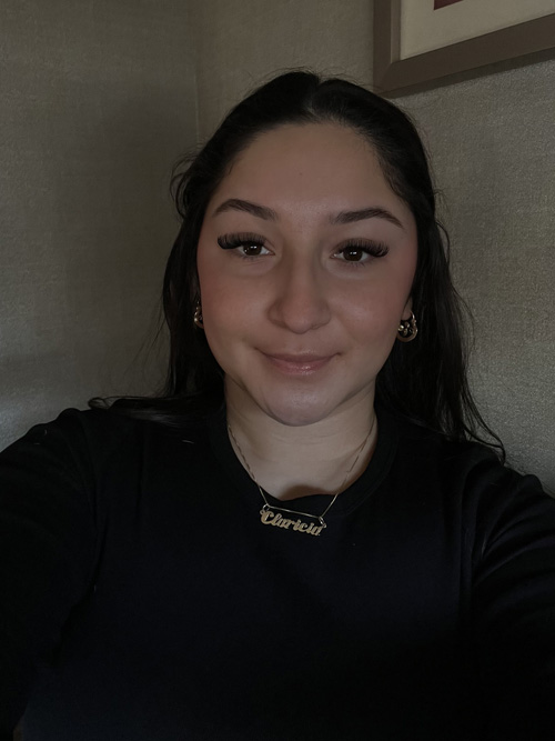 A woman with dark hair and gold jewelry smiles slightly in a dimly lit room.