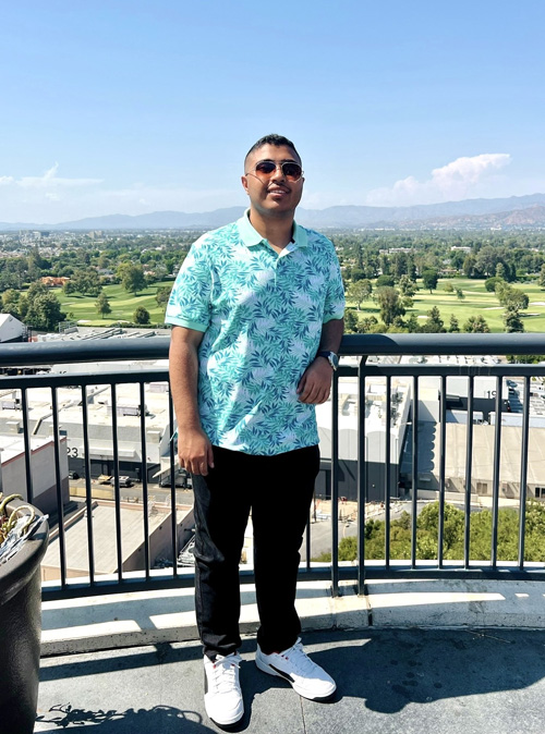 A man in sunglasses and a tropical shirt stands on a balcony overlooking a cityscape.
