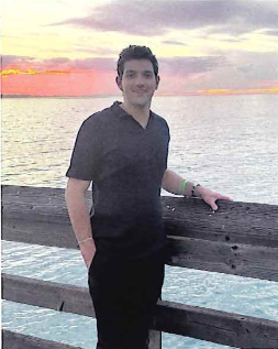 Amir Ayash standing by a wooden railing overlooking the ocean at sunset, wearing a dark shirt and smiling at the camera.