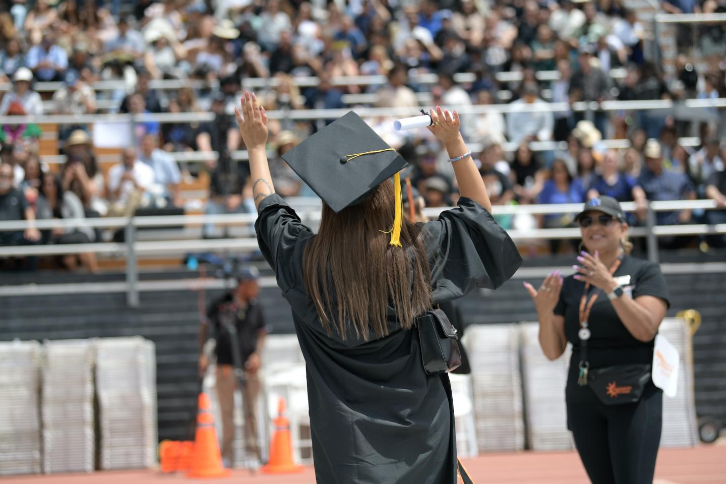 A graduate in cap and gown raises their arms while walking across the stage, with an applauding crowd in the stadium behind them.