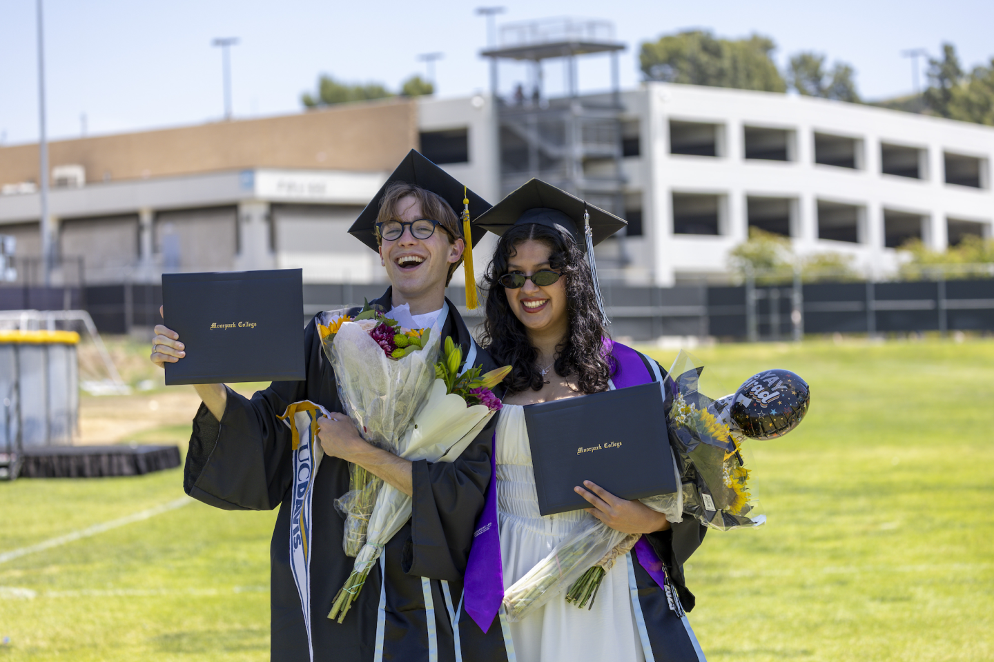 Two graduates in caps and gowns smile while holding diplomas and bouquets on a grassy field, with a campus building in the background.