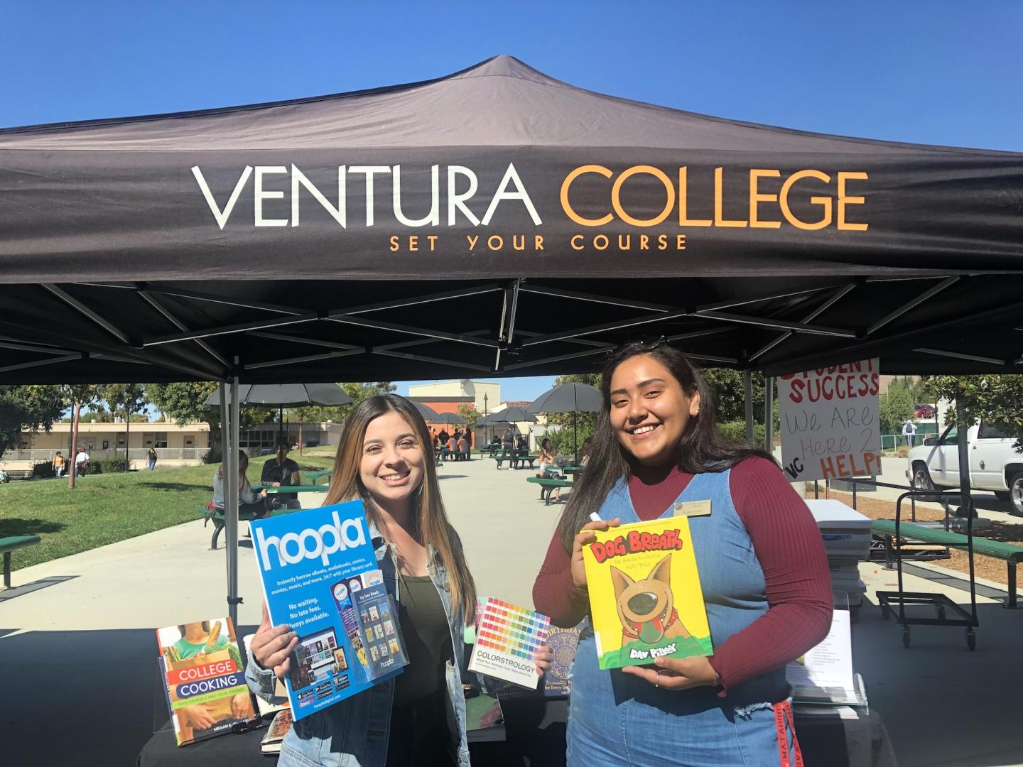 Two students smile while holding books at a Ventura College Student Success event under a branded canopy on campus.