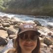 A person (Sarah Escobar) taking a selfie with a stream of water flowing over rocks surrounded by green trees. She is wearing a black Patagonia baseball hat and sunglasses.