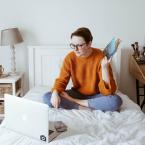 Student sitting on a bed with a laptop and studying a book.