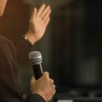 Photo of a person giving a speech while holding a microphone in one hand and gesturing with the other.