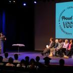 Photograph of Edgar Euan, a person wearing a black floral button down, giving the keynote speech at the VCCCD Lavender Graduation. On stage are 10 District employees and administrators, including the Chancellor and college presidents. Graduating students sit in the auditorium seats. An image that says "Proud to be VCCCD • Moorpark • Oxnard • Ventura • Community Colleges" in a circle is projected on the back of the stage. 