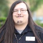 Photo of Alexander Rock Fredell, man with long brown hair and glasses wearing a black button down shirt, standing in front of greenery outdoors