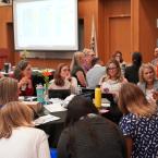 A group of educators and professionals are engaged in discussion at a Dual Enrollment Community of Practice event. They are seated at round tables, with laptops, notebooks, and drinks in front of them. A large presentation screen displaying charts is visible in the background, and the atmosphere is collaborative and focused.
