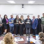 Classified Employee of the Year nominees stand holding plaques alongside the Chancellor and Board of Trustees in a boardroom. The group is smiling in front of a wall that reads “Service in the Public’s Interest.”