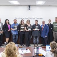 Classified Employee of the Year nominees stand holding plaques alongside the Chancellor and Board of Trustees in a boardroom. The group is smiling in front of a wall that reads “Service in the Public’s Interest.”