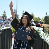 A graduate from Moorpark College wearing a cap and gown with a black stole waving at her family from the stage after accepting her diploma