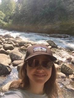 A person (Sarah Escobar) taking a selfie with a stream of water flowing over rocks surrounded by green trees. She is wearing a black Patagonia baseball hat and sunglasses.