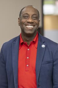 Dr. Julius Sokenu smiling, wearing a red shirt and blue spor Dr. Julius Sokenu smiling, wearing a red shirt and blue spor