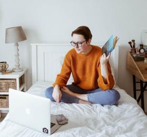 Student sitting on a bed with a laptop and studying a book.