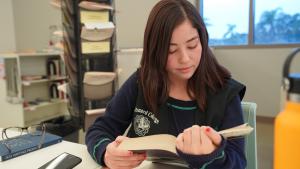 Young student wearing Oxnard College vest reading textbook in library.