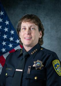 Police Chief Kelli Florman, white woman with short curly brown hair wearing her police uniform and sitting in front of an American flag