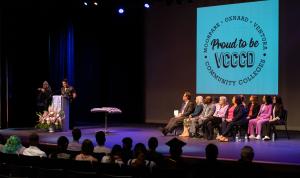 Photograph of Edgar Euan, a person wearing a black floral button down, giving the keynote speech at the VCCCD Lavender Graduation. On stage are 10 District employees and administrators, including the Chancellor and college presidents. Graduating students sit in the auditorium seats. An image that says "Proud to be VCCCD • Moorpark • Oxnard • Ventura • Community Colleges" in a circle is projected on the back of the stage. 