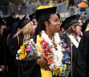 A smiling graduate holds a bouquet and wears colorful leis at the 2024 Ventura College Commencement Ceremony.