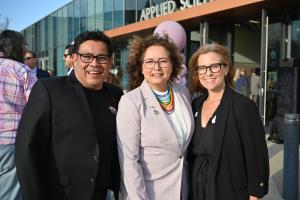 Ventura College President Claudia Lourido-Habib, center, stands smiling between event emcees Oscar Rivera, left, and Brook Masters, right, in front of the Applied Science Center at Ventura College during the 2025 Lavender Graduation. The group is outdoors on a sunny day, surrounded by attendees celebrating LGBTQIA+ graduates.