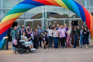 Students, faculty, and staff celebrate under a rainbow arch at Oxnard College's Lavender Graduation 2026, waving pride flags and cheering outside the campus entrance.