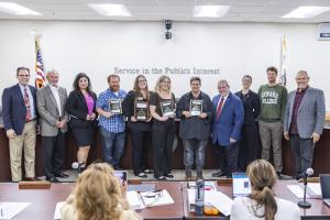 Classified Employee of the Year nominees stand holding plaques alongside the Chancellor and Board of Trustees in a boardroom. The group is smiling in front of a wall that reads “Service in the Public’s Interest.”